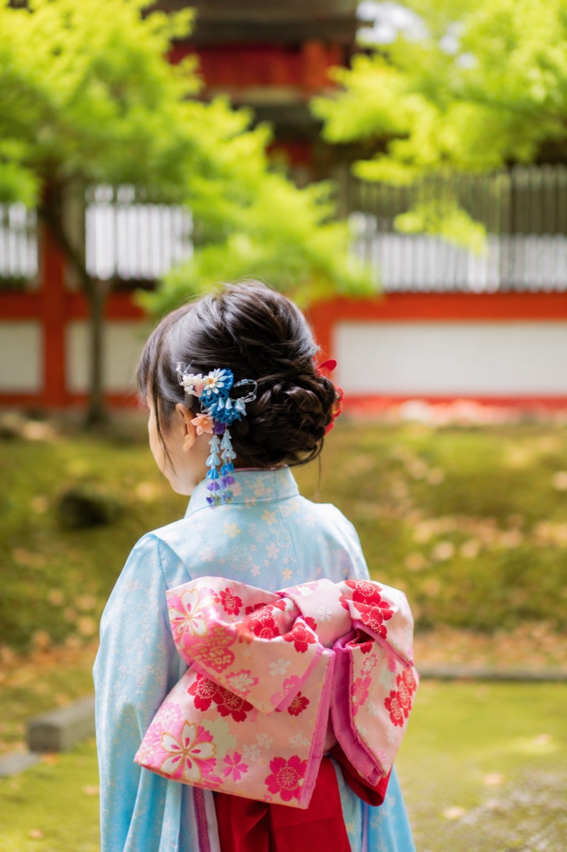 七五三の着物姿の女の子の後ろ姿。神社の朱塗りの塔を背景に